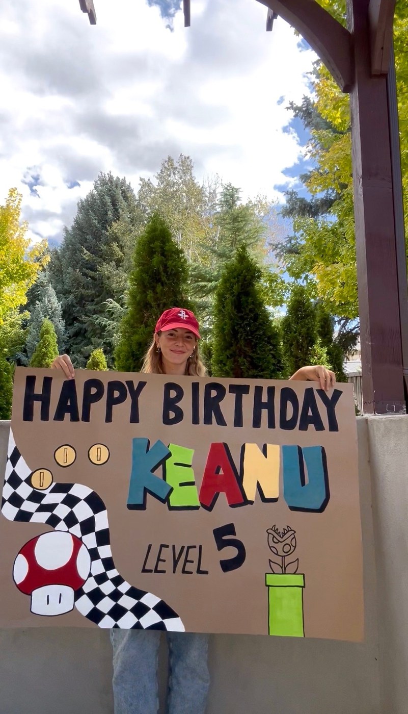 A woman holding a custom Mario-themed birthday banner that reads Happy Birthday Keanu, Level 5, with a mushroom, checkered flag and piranha plant